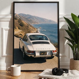May include: A framed wall art print featuring a white vintage Porsche 911 sports car parked on a coastal road overlooking the ocean and mountains. The car displays a California license plate reading CRM 168 and the Porsche logo on the rear deck.