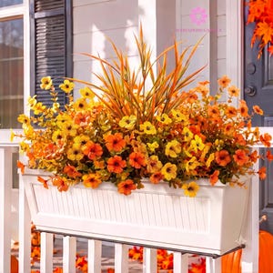 May include: A rectangular white planter box mounted on a railing, filled with an arrangement of artificial flowers in shades of yellow and orange, featuring tall, slender, pointed leaves in the center and small, five-petaled blossoms throughout.