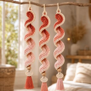 May include: Three hanging spiral decorations made of crocheted yarn in shades of red, coral, and pink. Each piece features a wooden bead, a small crocheted shell-shaped accent, and a dangling tassel at the bottom, suspended from a wooden branch.