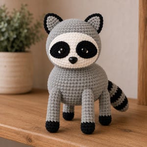 May include: A crochet raccoon toy featuring a gray body, a white face mask with black eye patches, black ears, and black paws. The raccoon has a round head, a small black nose, and a striped tail with alternating black and gray bands.