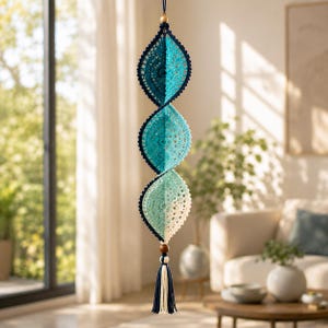 May include: A hanging mobile featuring three interconnected, leaf-shaped crochet ornaments in a gradient of dark blue, teal, and cream. The structure is suspended from a small wooden bead and terminates in a matching dark blue and cream tassel.