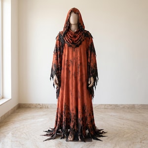 May include: A full-length hooded robe in a burnt orange hue featuring distressed, jagged edges along the hem and wide sleeves. The garment displays a dark, mottled black pattern throughout, with a draped cowl neckline and a pointed hood.