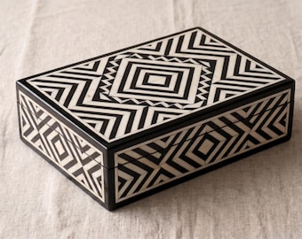May include: A rectangular box featuring a high-contrast black and white geometric pattern. The lid and sides are decorated with intricate, repeating chevron, diamond, and square motifs that create a bold, symmetrical visual design across the surface.