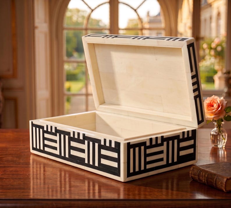 May include: A rectangular storage box with an open lid, featuring a cream-colored base with a contrasting black geometric pattern of vertical and horizontal lines. The interior is smooth and cream-colored, matching the exterior frame and lid.
