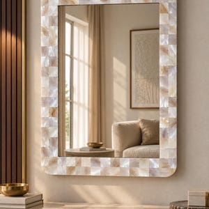 Op de afbeelding: Een rechthoekige wandspiegel met een frame bestaande uit kleine, vierkante, iriserende parelmoertegels in tinten crème, beige en zacht wit. De spiegel heeft afgeronde hoeken en een reflecterend glazen oppervlak dat een kamerinterieur toont.