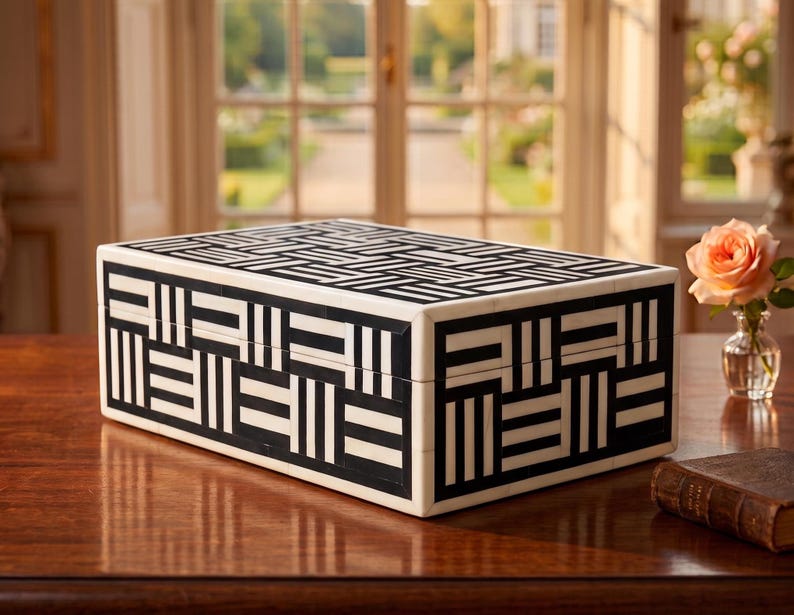 May include: A rectangular storage box featuring a high-contrast black and white geometric pattern. The exterior is decorated with an intricate, repeating arrangement of alternating horizontal and vertical stripes, creating a structured, woven-like visual effect.