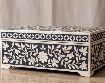 May include: A rectangular storage box featuring a black base with intricate white floral and leaf inlays. The lid is decorated with a repeating geometric circle pattern in black and white. The box has four small feet and a flat, hinged top surface.