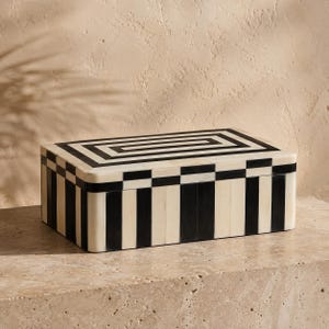 Op de afbeelding: Een rechthoekige opbergdoos met een contrastrijk zwart en crèmekleurig streeppatroon. Het deksel heeft een concentrisch rechthoekig ontwerp, terwijl de zijkanten zijn versierd met verticale strepen en een geblokte rand aan de bovenkant.