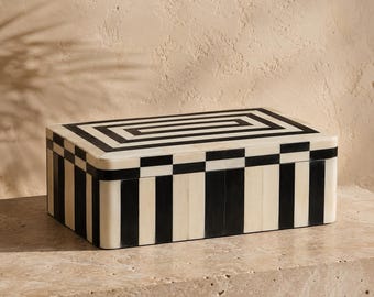 May include: A rectangular storage box featuring a high-contrast black and cream striped pattern. The lid displays a concentric rectangular design, while the sides are adorned with vertical stripes and a checkered border along the top edge.