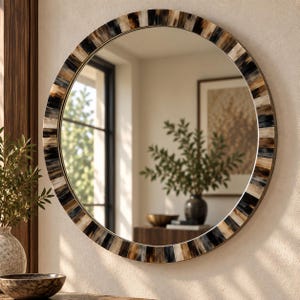 Puede incluir: Un gran espejo de pared redondo con un marco decorativo compuesto por segmentos verticales alternos en tonos negro, crema y marrón. La superficie de cristal reflectante muestra una vista clara de una habitación interior con una planta y muebles.