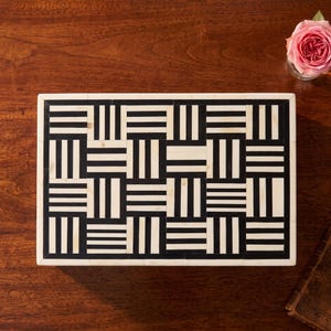 May include: A rectangular box featuring a geometric pattern of alternating black and off-white stripes arranged in a basket-weave style. The lid is framed by a solid off-white border, creating a high-contrast, repeating grid design across the surface.