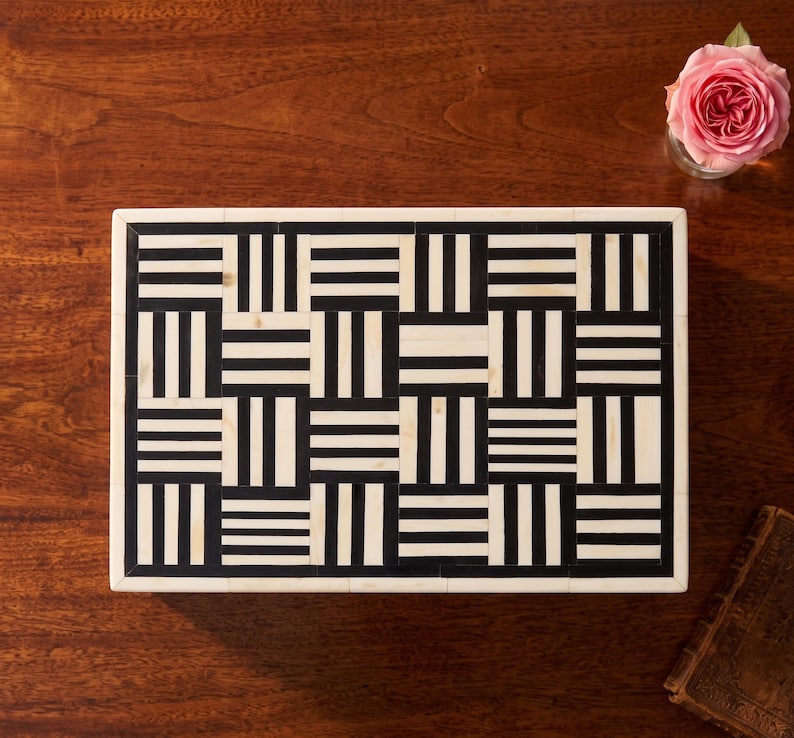 May include: A rectangular box featuring a geometric pattern of alternating black and off-white stripes arranged in a basket-weave style. The lid is framed by a solid off-white border, creating a high-contrast, repeating grid design across the surface.