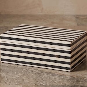 May include: A rectangular storage box featuring a high-contrast pattern of alternating horizontal black and off-white stripes. The box has a flat lid and straight edges, with the striped design continuing seamlessly across the top and sides.