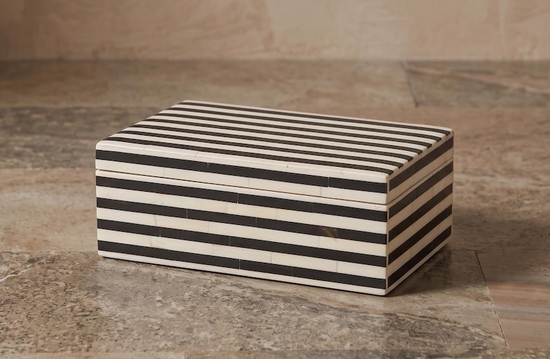 May include: A rectangular storage box featuring a high-contrast pattern of alternating horizontal black and off-white stripes. The box has a flat lid and straight edges, with the striped design continuing seamlessly across the top and sides.