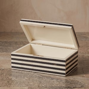 May include: A rectangular storage box with a hinged lid, featuring a repeating pattern of horizontal black and off-white stripes. The interior of the box is smooth and off-white, with visible metal hinges connecting the lid to the base.