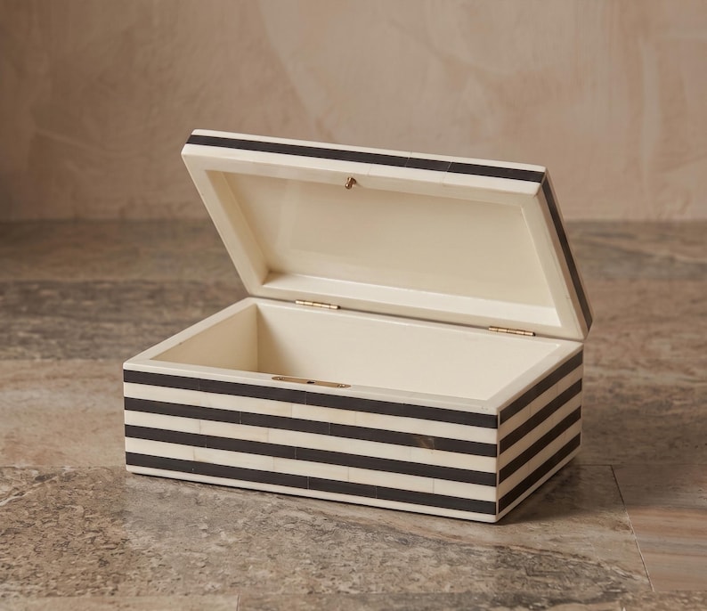 May include: A rectangular storage box with a hinged lid, featuring a repeating pattern of horizontal black and off-white stripes. The interior of the box is smooth and off-white, with visible metal hinges connecting the lid to the base.