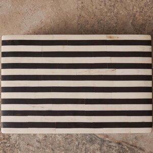 May include: A rectangular box featuring a high-contrast pattern of horizontal black and off-white stripes. The surface is composed of numerous small, rectangular segments arranged in a repeating linear design across the entire top face.