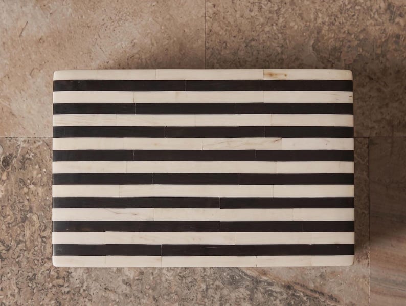 May include: A rectangular box featuring a high-contrast pattern of horizontal black and off-white stripes. The surface is composed of numerous small, rectangular segments arranged in a repeating linear design across the entire top face.