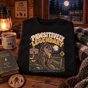 May include: A black sweatshirt featuring a graphic print with the text PAWSITIVELY LEGENDARY. The illustration depicts a furry creature eating a piece of cheese near a sign that reads BRAY ROAD, with the text BEAST OF BRAY ROAD ESTABLISHED 1936.
