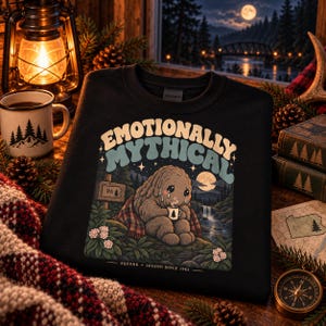 May include: A black sweatshirt featuring a graphic of a crying, furry creature holding a small mug. The text reads "EMOTIONALLY MYTHICAL" and "SQUONK LEGEND SINCE 1963". The illustration includes a forest, a waterfall, a moon, and a sign labeled "PA".