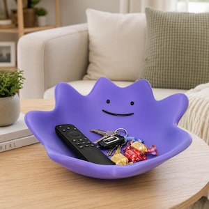 May include: A purple, amorphous-shaped bowl featuring a simple, minimalist face with two black dot eyes and a thin, curved black line for a mouth. The bowl contains a black remote control, a set of metal keys, and several colourful wrapped sweets.