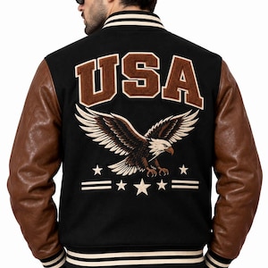 Op de afbeelding: Een zwart varsity-jack met bruine mouwen, voorzien van een grote geborduurde adelaarsafbeelding op de achterkant boven de tekst USA. Het jack heeft gestreepte geribbelde boorden bij de kraag, mouwen en zoom, met extra ster- en lijnstiksels.