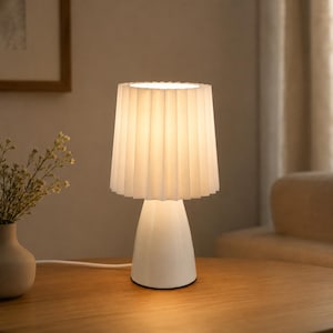 Könnte beinhalten: Eine weiße Tischleuchte mit einem plissierten zylindrischen Lampenschirm und einem glatten, konischen Fuß. Die Lampe strahlt ein warmes, sanftes Licht aus, das die Umgebung erhellt. Das Design ist schlicht und einfach, mit einem weißen Netzkabel.