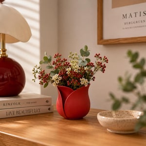 May include: A red ceramic vase shaped like a tulip blossom, containing a bouquet of small white flowers, red berries, and green eucalyptus leaves. The vase has a smooth, matte finish and curved petals that form the opening for the floral arrangement.