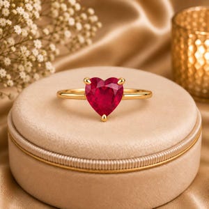 May include: A gold ring featuring a single heart-shaped red gemstone set in a four-prong setting. The band is thin and polished, showcasing a smooth metallic finish that complements the vibrant red color of the faceted heart-shaped stone.