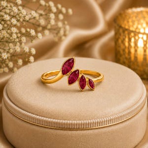 May include: A gold ring featuring four marquise-cut deep red gemstones arranged in a graduated, leaf-like pattern along the top of the band. The stones are set in polished gold metal, creating a curved, asymmetrical design that follows the finger.