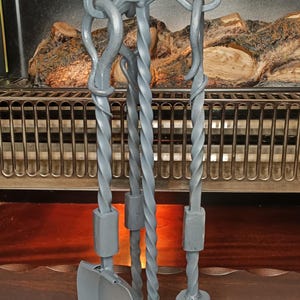 May include: A matte grey metal fireplace tool set featuring a shovel, a poker, and a brush. Each tool has a twisted handle design and hangs from a central stand with a three-legged base. The set includes a brush with dark bristles.