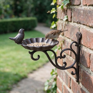 May include: A dark brown cast iron wall-mounted bird feeder featuring a decorative scrollwork bracket and a scalloped bowl filled with mixed birdseed. A small brown sparrow stands inside the bowl, while a stationary bird figurine sits on the edge.