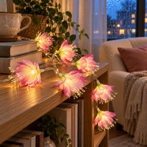 May include: A string of LED fairy lights featuring pink, flower-shaped covers with thin, translucent petals and glowing centers. The lights are draped across a wooden surface, resting near a stack of books and a ceramic mug.