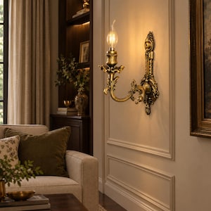 May include: A wall-mounted sconce featuring an ornate, antique gold-toned metal finish with intricate scrollwork and leaf-like detailing. The fixture holds a single upright candle-style light bulb that emits a warm, soft glow against the wall.
