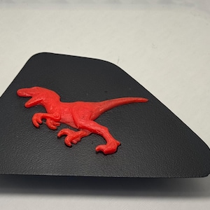 May include: A black, triangular-shaped plastic component featuring a raised, bright red dinosaur figure in the center. The dinosaur is depicted in a side profile, showing a raptor-like creature with sharp claws and a long tail, textured onto the surface.