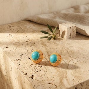 May include: A pair of gold-toned stud earrings featuring a central round turquoise stone. The gold metal surface has a textured, granular finish with small embedded green accents. The earrings are circular in shape and display a detailed, ornate metalwork pattern surrounding the vibrant blue-green stone.
