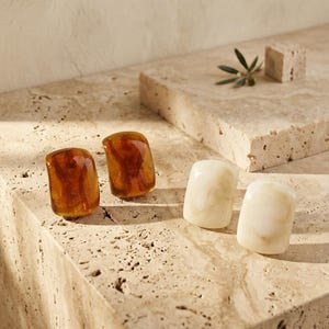 May include: Two pairs of rectangular stud earrings. One pair is made of translucent amber-colored resin, while the other pair is made of opaque, marbled off-white resin. Each earring has a smooth, rounded surface and a polished finish.