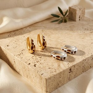 May include: Two pairs of hoop earrings, one pair in gold-tone metal and one pair in silver-tone metal. Each hoop features a series of recessed, dark brown, organic-shaped cutouts along the outer surface, creating a textured, porous appearance.
