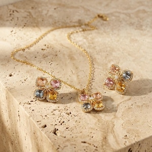 May include: A gold-tone necklace and matching earrings set featuring four-leaf clover motifs. Each clover is composed of four cushion-cut multicolored gemstones in pink, blue, and yellow, accented with small clear crystals and a central star detail.