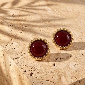 May include: A pair of round stud earrings featuring a smooth, deep burgundy cabochon center surrounded by a polished gold-tone twisted rope border. The earrings have a circular shape and a reflective finish on both the stone and the metal frame.