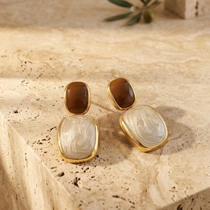 Può includere: Un paio di orecchini color oro con un design a due parti. Ogni orecchino presenta una piccola parte superiore ovale di colore marrone scuro liscio, collegata a una parte inferiore rettangolare più grande con una finitura smaltata bianco perla e crema.