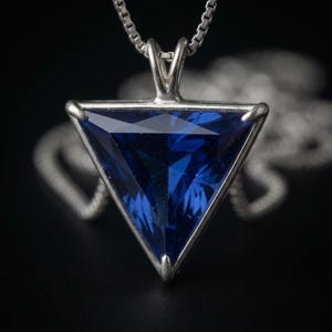 May include: A silver necklace featuring a triangular pendant with a faceted deep blue gemstone. The pendant is held by a silver prong setting and hangs from a delicate silver chain, showcasing a clear geometric shape and a polished metallic finish.