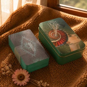May include: A deck of tarot cards with dark covers featuring teal starburst designs and a matching green rectangular metal tin. The tin lid displays the text The Light Seer's Tarot and an illustration of a person with a red spiral headpiece.