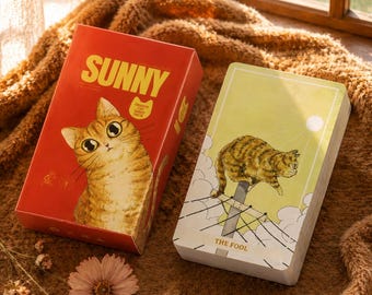 May include: A red rectangular box labeled SUNNY ORANGE CAT TAROT DECK with an illustration of an orange tabby cat, alongside a deck of cards featuring a light green card titled THE FOOL showing an orange cat perched on a utility pole wire.