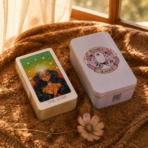 Op de afbeelding: Een set tarotkaarten met de kaart De Ster bovenop, voorzien van een kleurrijke illustratie van een persoon met lang donker haar en een stralende ster, naast een wit metalen opbergblik met de tekst Britt's 3rd Eye Tarot.