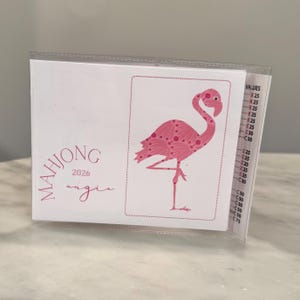 May include: A pink flamingo illustration on a white card, part of a Mahjong set. The card has the text "MAHJONG 2026 angie". The flamingo is in a standing pose with a patterned design. The card is in a clear plastic holder.