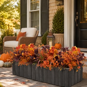 May include: A rectangular dark gray wooden planter box filled with a dense arrangement of artificial autumn foliage, including orange, yellow, and deep red maple leaves, small orange flowers, and thin green grasses, creating a vibrant seasonal display.