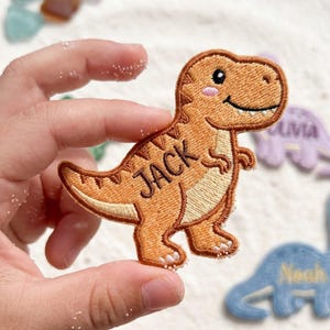 May include: An embroidered iron-on patch shaped like a brown Tyrannosaurus Rex dinosaur. The dinosaur features a cream-colored belly, a small pink cheek detail, and the name JACK stitched in dark brown thread across its side body.