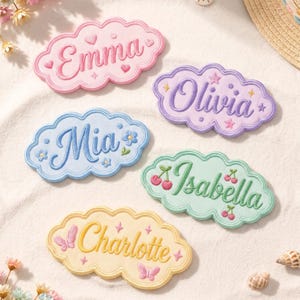 May include: Five cloud-shaped embroidered patches with names in cursive script. The pink patch says Emma with hearts, purple says Olivia with stars, blue says Mia with flowers, green says Isabella with cherries, and yellow says Charlotte with butterflies.