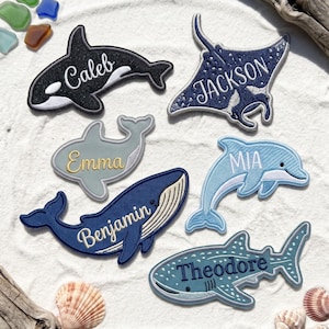 May include: Six embroidered fabric patches shaped like sea creatures, including an orca labeled Caleb, a stingray labeled Jackson, a shark labeled Emma, a dolphin labeled Mia, a whale labeled Benjamin, and a whale shark labeled Theodore.